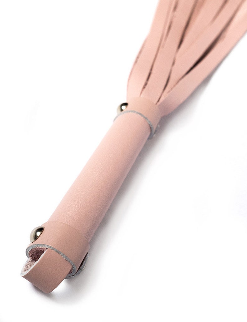 A close-up of the handle of the Stupid Cute Baby Pink Leather Flogger is shown against a blank background, displaying the small leather loop at the base of the handle and the metal hardware.