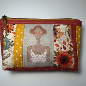 May include: A quilted cosmetic bag with a zipper closure. The bag features a central panel with an illustration of a woman in a floral dress. The bag's design incorporates floral and polka dot fabric panels in shades of orange, yellow, and cream.
