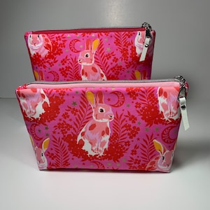 May include: Two pink zippered pouches with a repeating pattern of pink bunnies, crescent moons, and gold stars on a pink background.