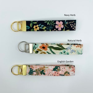May include: Three fabric keychains with gold and silver key rings. The keychains feature floral patterns in navy, cream, and pink. The text "Navy Herb", "Natural Herb", and "English Garden" is printed above each keychain.