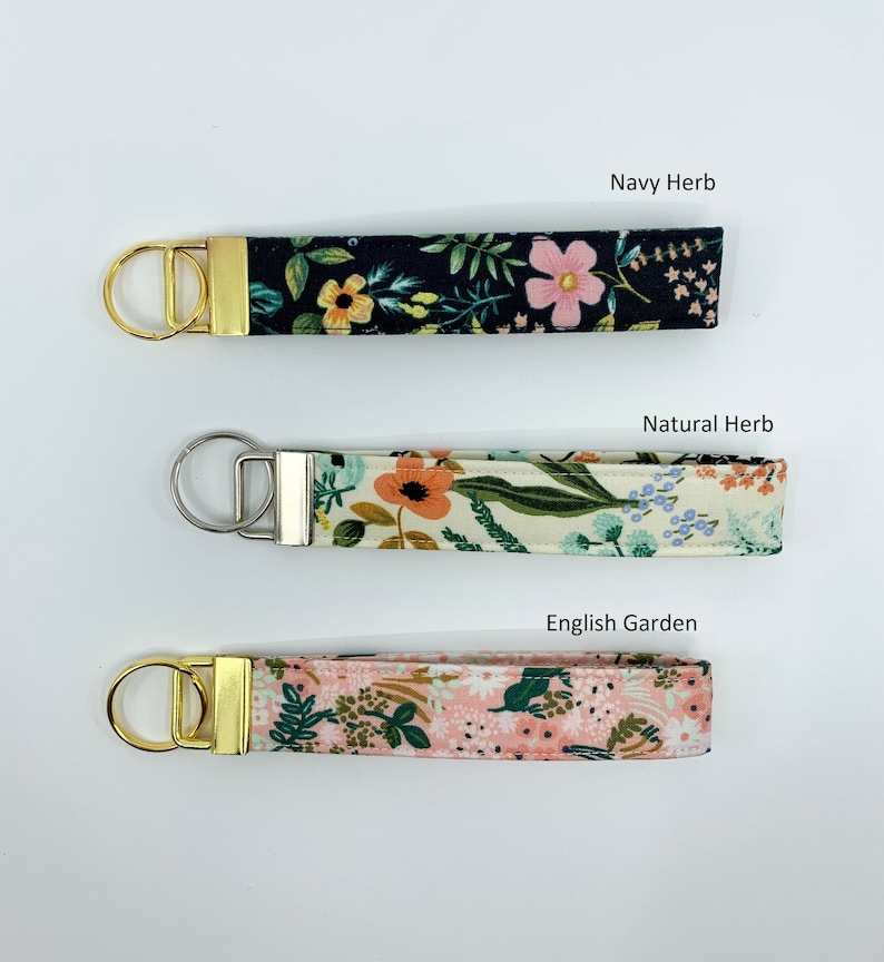 May include: Three fabric keychains with gold and silver key rings. The keychains feature floral patterns in navy, cream, and pink. The text "Navy Herb", "Natural Herb", and "English Garden" is printed above each keychain.