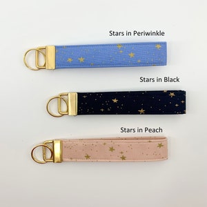 May include: Three keychains with gold key rings and fabric straps. The straps are printed with gold stars on a blue, black, and peach background. The text "Stars in Periwinkle", "Stars in Black", and "Stars in Peach" is printed above each keychain.