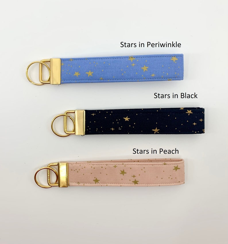 May include: Three keychains with gold key rings and fabric straps. The straps are printed with gold stars on a blue, black, and peach background. The text "Stars in Periwinkle", "Stars in Black", and "Stars in Peach" is printed above each keychain.