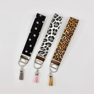 May include: Three keychains with different patterned fabric straps and silver key rings. The straps are black with white paw prints, gray with black leopard print, and brown with orange leopard print. Each keychain has a tassel attached to the key ring. The tassels are gray, pink, and brown.