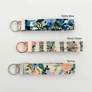 May include: Three keychains with different fabric patterns. The top keychain has a blue floral pattern and the text "Petite Blue". The middle keychain has a black and white music note pattern and the text "Music Notes". The bottom keychain has a floral pattern and the text "Spring".
