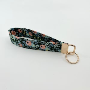 May include: A green fabric key chain with a floral pattern featuring pink and blue flowers. The key chain has a gold metal key ring.