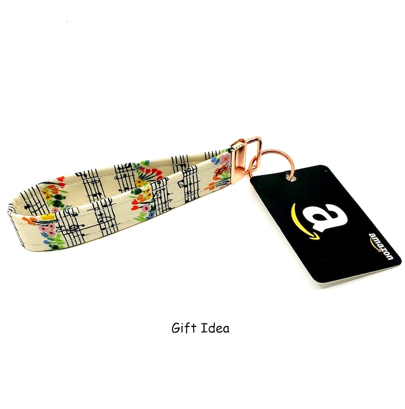 May include: A white and cream key chain with a musical note and floral pattern. The key chain has a rose gold metal ring and clasp. The text "Gift Idea" is printed below the key chain.