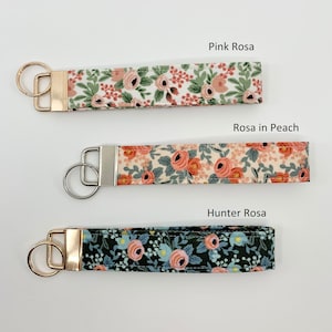 May include: Three keychains with floral patterns. The top keychain is white with pink flowers and green leaves and has the text "Pink Rosa". The middle keychain is cream with pink flowers and green leaves and has the text "Rosa in Peach". The bottom keychain is dark green with pink flowers and blue leaves and has the text "Hunter Rosa".