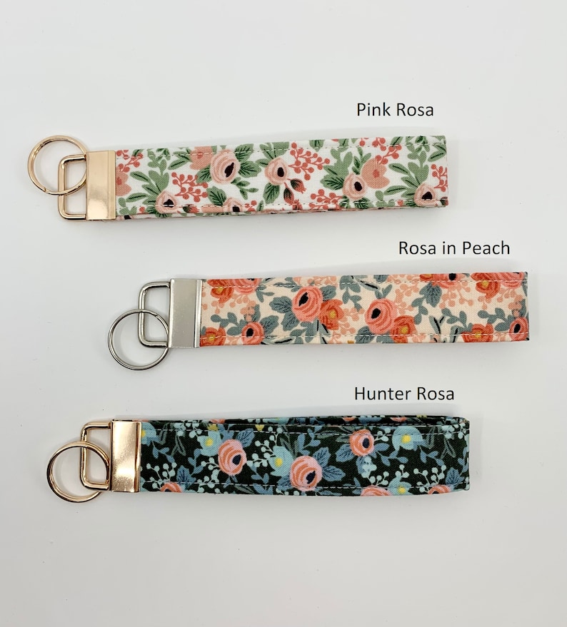 May include: Three keychains with floral patterns. The top keychain is white with pink flowers and green leaves and has the text "Pink Rosa". The middle keychain is cream with pink flowers and green leaves and has the text "Rosa in Peach". The bottom keychain is dark green with pink flowers and blue leaves and has the text "Hunter Rosa".