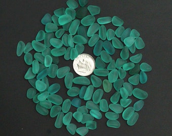 Op de afbeelding: Een grote verzameling turquoise zeeglasstukken, sommige met een meer ondoorzichtige afwerking, gerangschikt in een cirkelvormige patroon op een zwarte achtergrond. Een Nederlandse 10-cent munt is opgenomen voor schaal.
