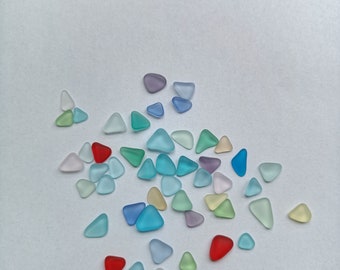 May include: A collection of small, triangular pieces of sea glass in various shades of blue, green, red, and purple. The sea glass is scattered on a white surface.