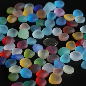 May include: A collection of colourful, frosted glass pebbles in various shades of blue, green, red, pink, and yellow. The pebbles are scattered on a black surface.