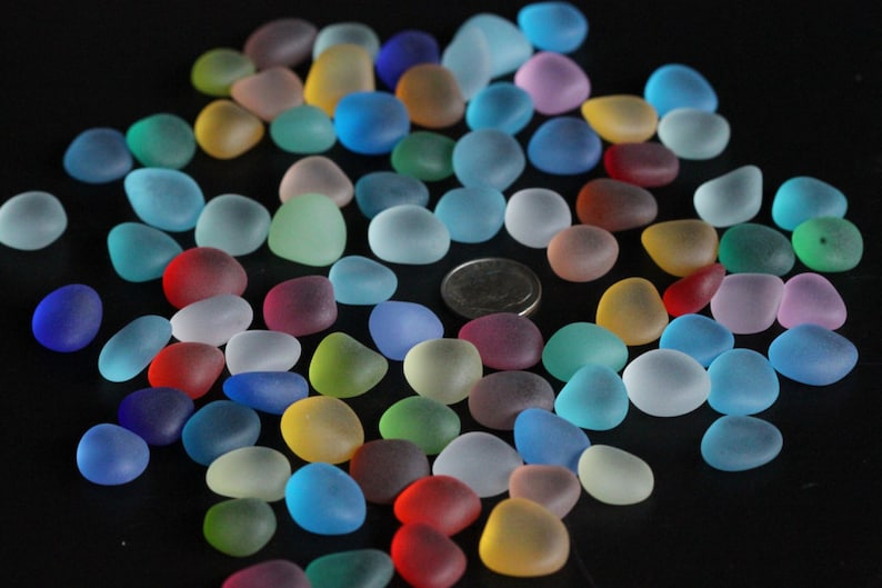 May include: A collection of colourful, frosted glass pebbles in various shades of blue, green, red, pink, and yellow. The pebbles are scattered on a black surface.