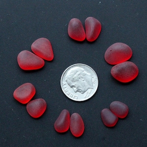 Pode incluir: Uma coleção de dez peças de vidro do mar vermelho, cada uma com a forma de uma lágrima, dispostas em círculo em torno de uma moeda de dez centavos de dólar americano de prata. A moeda é datada de 1968 e diz "LIBERTY" e "IN GOD WE TRUST".
