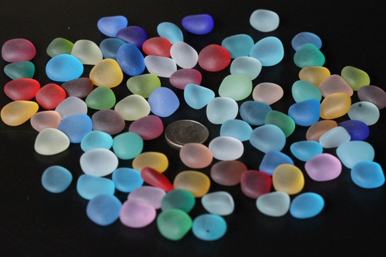 May include: A collection of colourful, frosted glass pebbles in various shades of blue, green, red, pink, and yellow. The pebbles are scattered on a black surface.