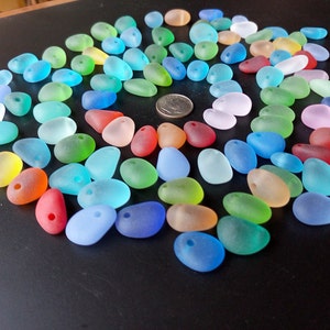 May include: A collection of colorful, frosted glass beads in various shades of blue, green, red, yellow, and pink. The beads are shaped like pebbles and have a hole drilled through them. They are arranged in a circle on a black surface.