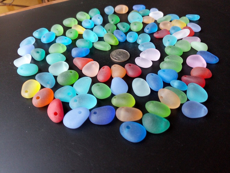 May include: A collection of colorful, frosted glass beads in various shades of blue, green, red, yellow, and pink. The beads are shaped like pebbles and have a hole drilled through them. They are arranged in a circle on a black surface.