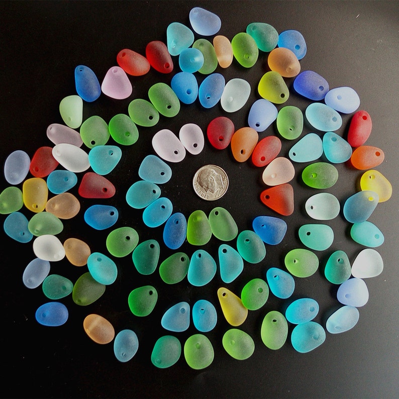 May include: A collection of colorful, frosted glass sea glass pieces with a hole drilled through each one. The sea glass is arranged in a circle on a black background.