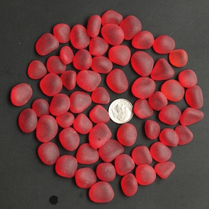 Puede incluir: Una colección de aproximadamente 40 piedras de vidrio rojo, algunas con un acabado esmerilado, dispuestas en círculo sobre una superficie negra. Se incluye una moneda de veinticinco centavos de dólar estadounidense de plata para la escala.