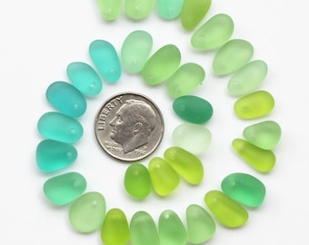 May include: A collection of 20 smooth, sea glass beads in various shades of green and blue. The beads are arranged in a semi-circle, with a ten pence coin for scale.