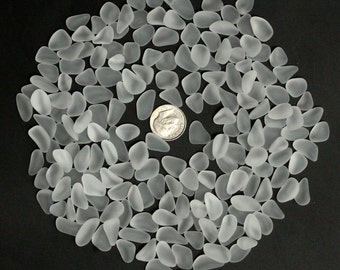May include: A collection of frosted, translucent sea glass pebbles in various shapes and sizes, arranged in a circular pattern. A silver dime is in the center for size comparison. The sea glass has a soft, matte finish, and the background is black.