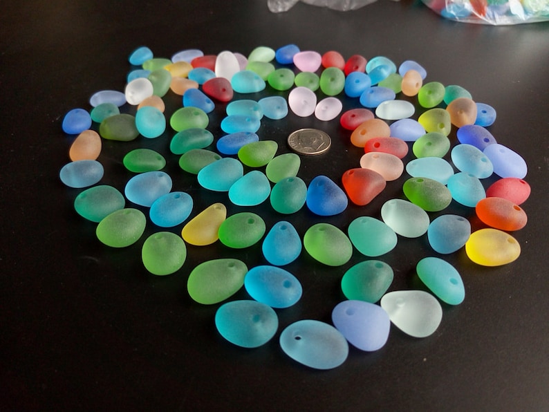 May include: Assortment of colorful, frosted glass beads in various shades of blue, green, red, and yellow. The beads are oval shaped and have a hole drilled through the top for stringing.