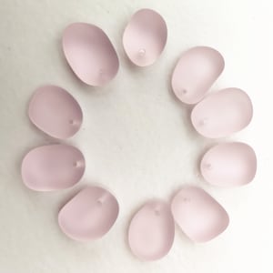 Puede incluir: Diez cuentas de vidrio de color rosa pálido con un agujero en el centro de cada cuenta. Las cuentas están dispuestas en círculo sobre una superficie blanca.