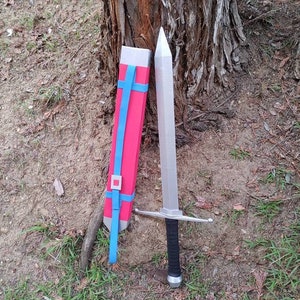 Puede incluir: Una espada plateada con empuñadura y guarda negras descansa en el suelo junto a una vaina roja con correas azules. La espada está cerca de un tronco de árbol, con hierba y tierra en el fondo.