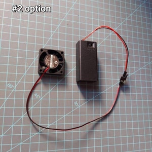 May include: A black computer fan with red and black wires connected to a black battery pack. The battery pack has a switch on the side. The fan and battery pack are on a gray grid surface. #2 option