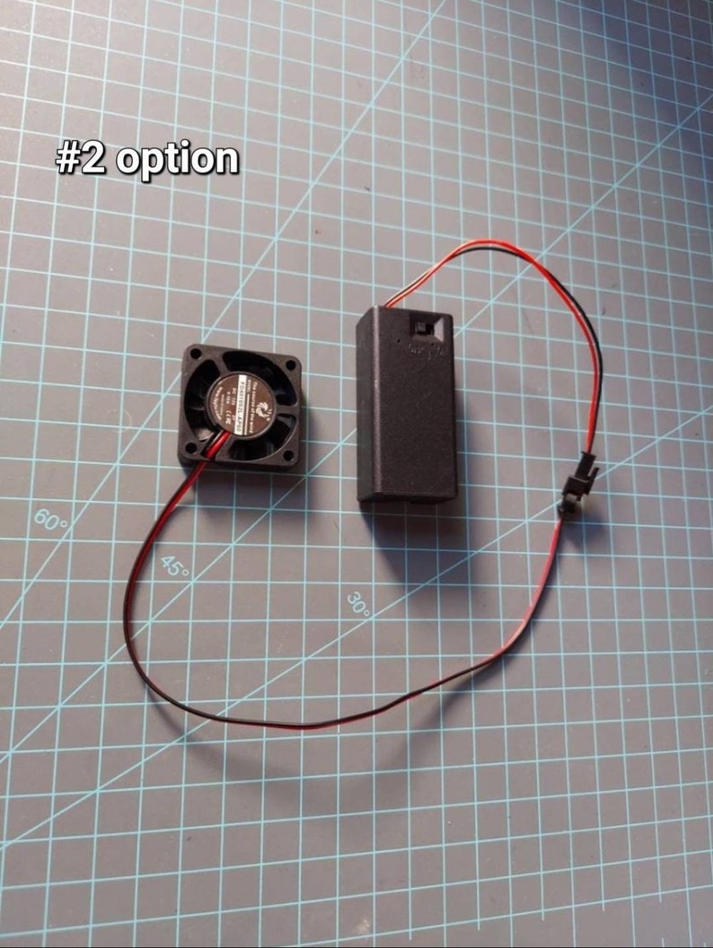 May include: A black computer fan with red and black wires connected to a black battery pack. The battery pack has a switch on the side. The fan and battery pack are on a gray grid surface. #2 option