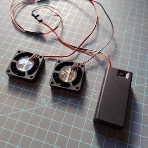 May include: Two black computer fans with red and black wires connected to a black battery pack. The fans are on a gray grid surface.