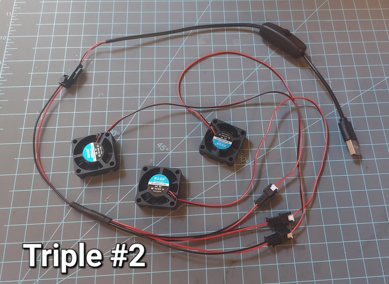 May include: A set of three small black fans with blue and white labels, connected by red and black wires. The wires lead to a USB connector and a switch. The text "Triple #2" is visible.