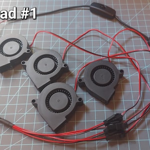 May include: Four black fans with red and black wires, connected to a USB plug and a switch. The text "Quad #1" is in the upper left corner. The fans are arranged on a gray surface.