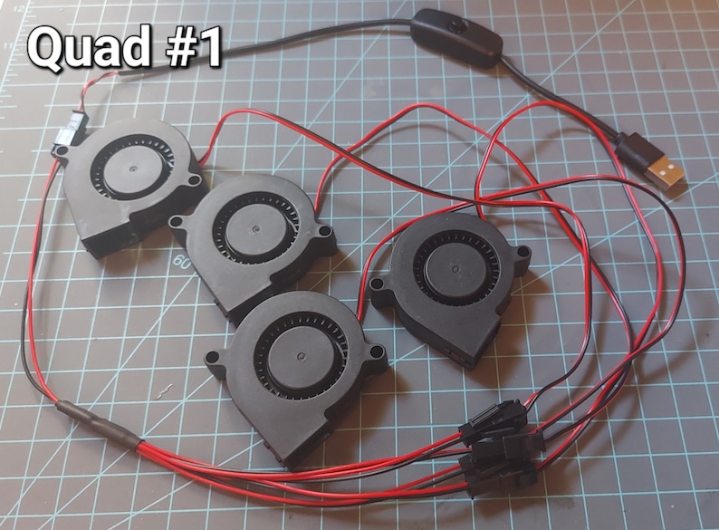 May include: Four black fans with red and black wires, connected to a USB plug and a switch. The text "Quad #1" is in the upper left corner. The fans are arranged on a gray surface.