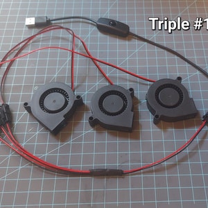 May include: Three black fan units with red and black wiring, connected to a USB cable and a switch. The text "Triple #1" is visible in the upper right corner. The fans are arranged on a gray grid surface.