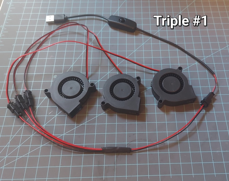 May include: Three black fan units with red and black wiring, connected to a USB cable and a switch. The text "Triple #1" is visible in the upper right corner. The fans are arranged on a gray grid surface.