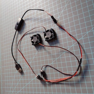 May include: Two small black fans with red and black wires, a USB connector, and a black on/off switch. The components are arranged on a gray grid surface, suggesting a DIY or electronics project.