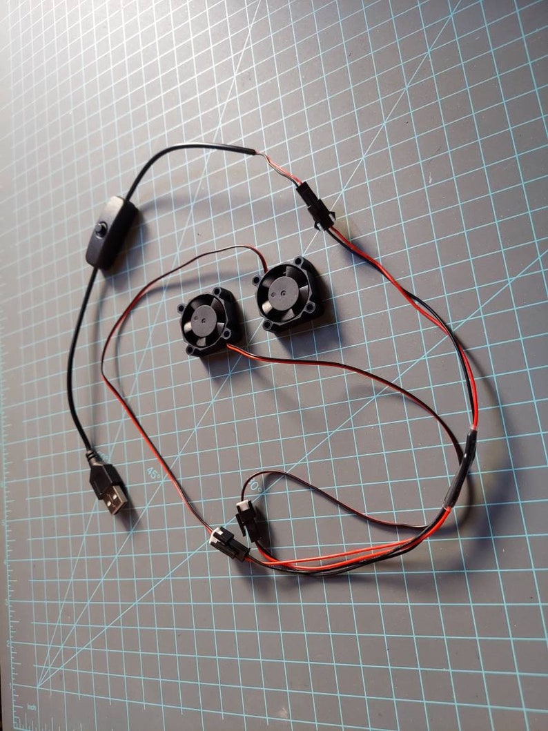 May include: Two small black fans with red and black wires, a USB connector, and a black on/off switch. The components are arranged on a gray grid surface, suggesting a DIY or electronics project.