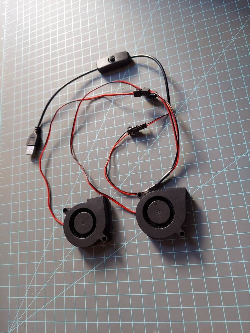 May include: Two small black fans with red and black wires, connected to a USB cable and a switch. The fans are on a gray grid surface.