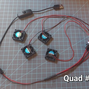 May include: An overhead shot shows a DIY electronic component setup. Four small black fans with blue and white labels are connected with red and black wires. A black USB connector, a switch, and a connector are also visible. The text "Quad #2" is in the bottom right.