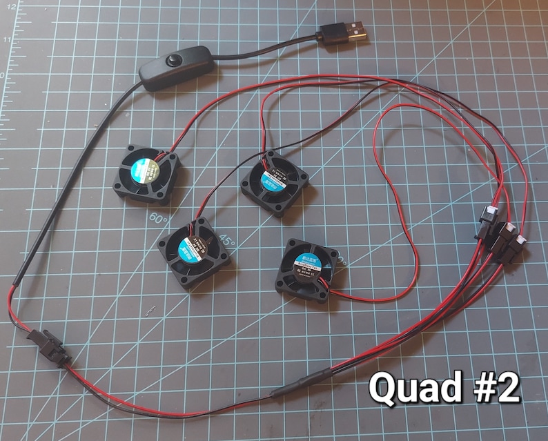 May include: An overhead shot shows a DIY electronic component setup. Four small black fans with blue and white labels are connected with red and black wires. A black USB connector, a switch, and a connector are also visible. The text "Quad #2" is in the bottom right.