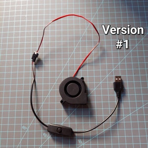 May include: A black USB-powered fan with a red and black wire, a black on/off switch, and the text "Version #1". The fan is circular with a central intake and a USB connector.