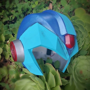 Puede incluir: Un casco azul y plateado con una visera roja. El casco está hecho de plástico y tiene una superficie texturizada. Está sentado en un lecho de plantas verdes.