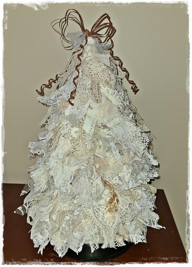 May include: A decorative Christmas tree made of layered white and cream-colored lace and fabric. The tree is topped with a brown ribbon bow and curly accents. The tree is displayed on a dark brown surface.