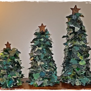 May include: Three handmade Christmas trees of varying sizes. Each tree is crafted from green fabric scraps, creating a textured, layered effect. They are topped with rustic, star-shaped ornaments. The trees are arranged on a wooden surface.
