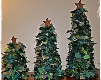May include: Three handmade Christmas trees of varying sizes. Each tree is crafted from green fabric scraps, creating a textured, layered effect. They are topped with rustic, star-shaped ornaments. The trees are arranged on a wooden surface.