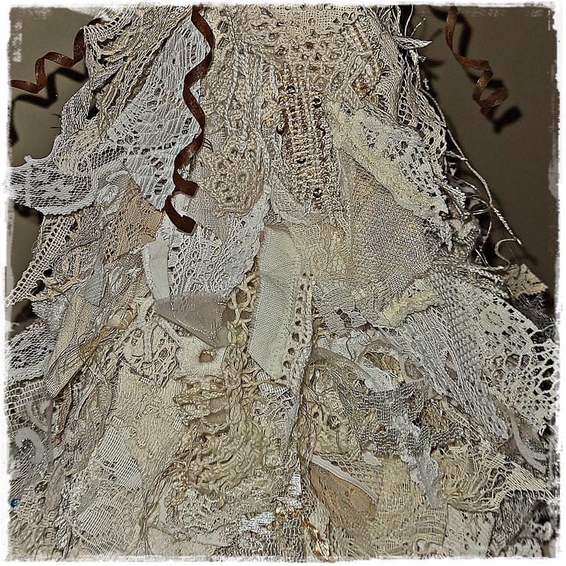 May include: Close-up of a handmade, layered textile art piece. The artwork features a variety of off-white and cream-colored lace, fabric scraps, and ribbons. The textures and patterns create a unique, vintage-inspired aesthetic. Brown ribbon accents add a touch of contrast.