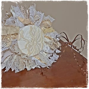 May include: A handmade, layered fabric flower in shades of white and cream, with intricate lace details. The flower is centered on a wooden surface and has a brown ribbon tied in a bow.
