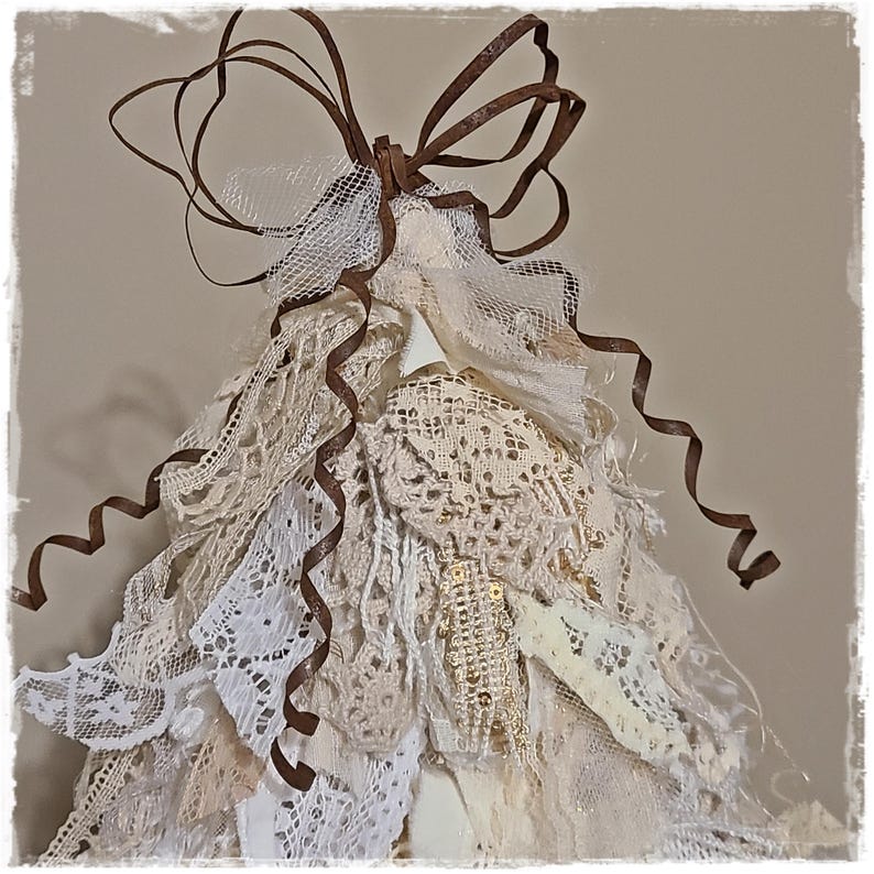 May include: A handmade, layered tree ornament with a rustic, vintage aesthetic. The tree is constructed from various lace fabrics in shades of white and cream, accented with brown ribbon and a decorative bow at the top. The ornament has a whimsical, handcrafted appearance.