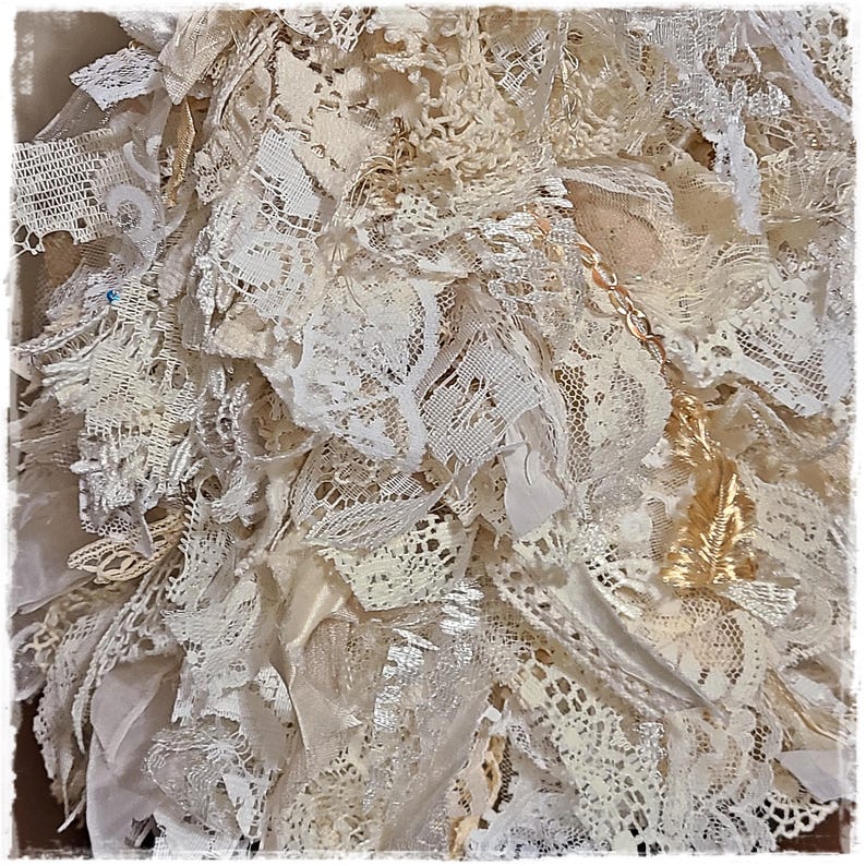 May include: Close-up of a textured, layered artwork composed of various off-white and cream-colored lace and fabric scraps. The composition includes different lace patterns, ribbons, and textures, creating a tactile and visually rich surface. A gold chain is visible.